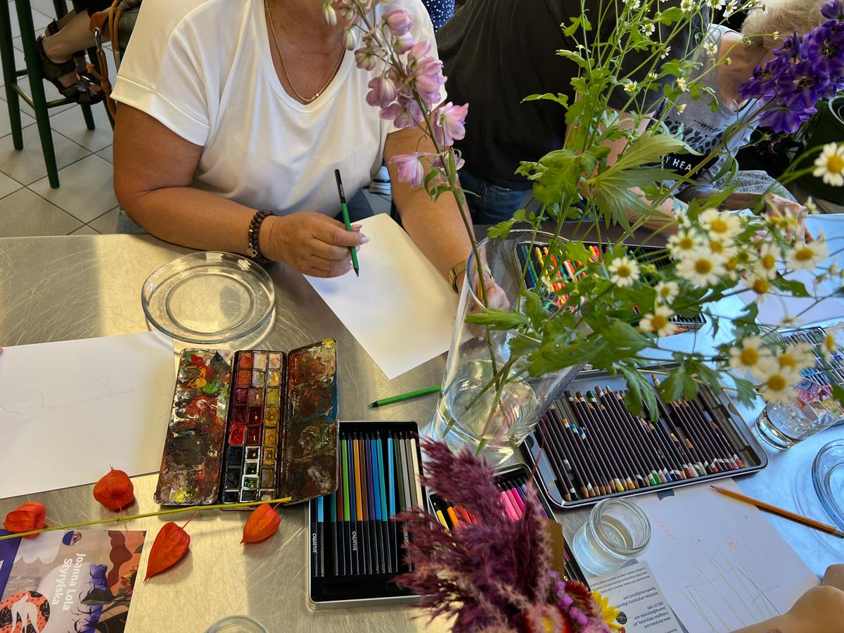 Kobieta w białej koszulce rysuje przy stole pełnym kolorowych pisaków, akwareli i kwiatów w szklankach.