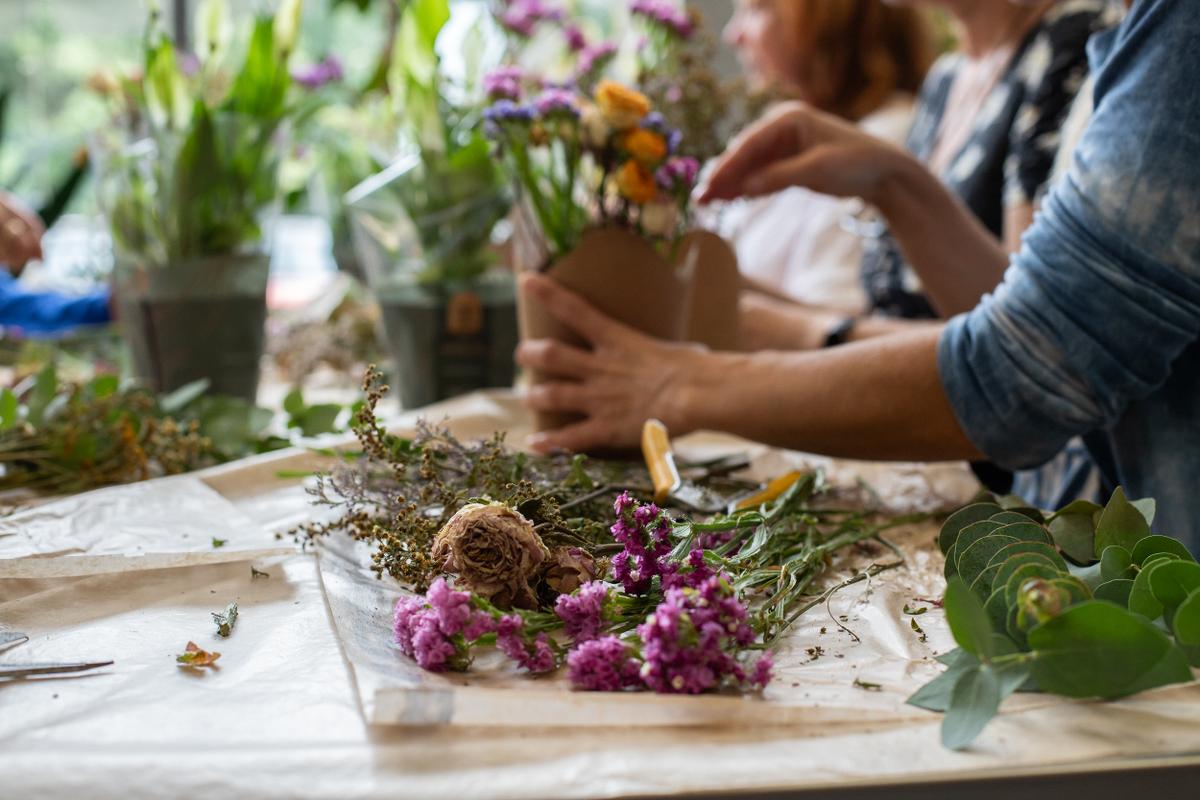 Grupa osób przygotowuje kwitnące kompozycje florystyczne przy drewnianym stole, pracując z kolorowymi kwiatami i zielenią.