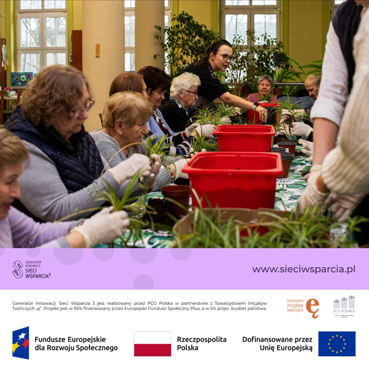 Grupa osób uczestniczy w warsztatach ogrodniczych, sadzając rośliny w czerwonych doniczkach w hali wypełnionej zielenią.