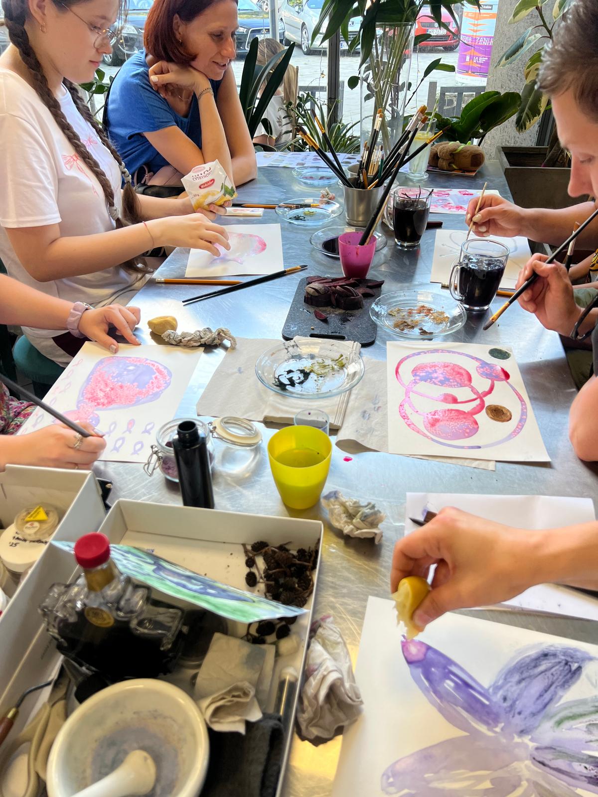 Grupa osób uczestniczy w warsztacie artystycznym, malując przy stole wśród roślin i materiałów do twórczości.