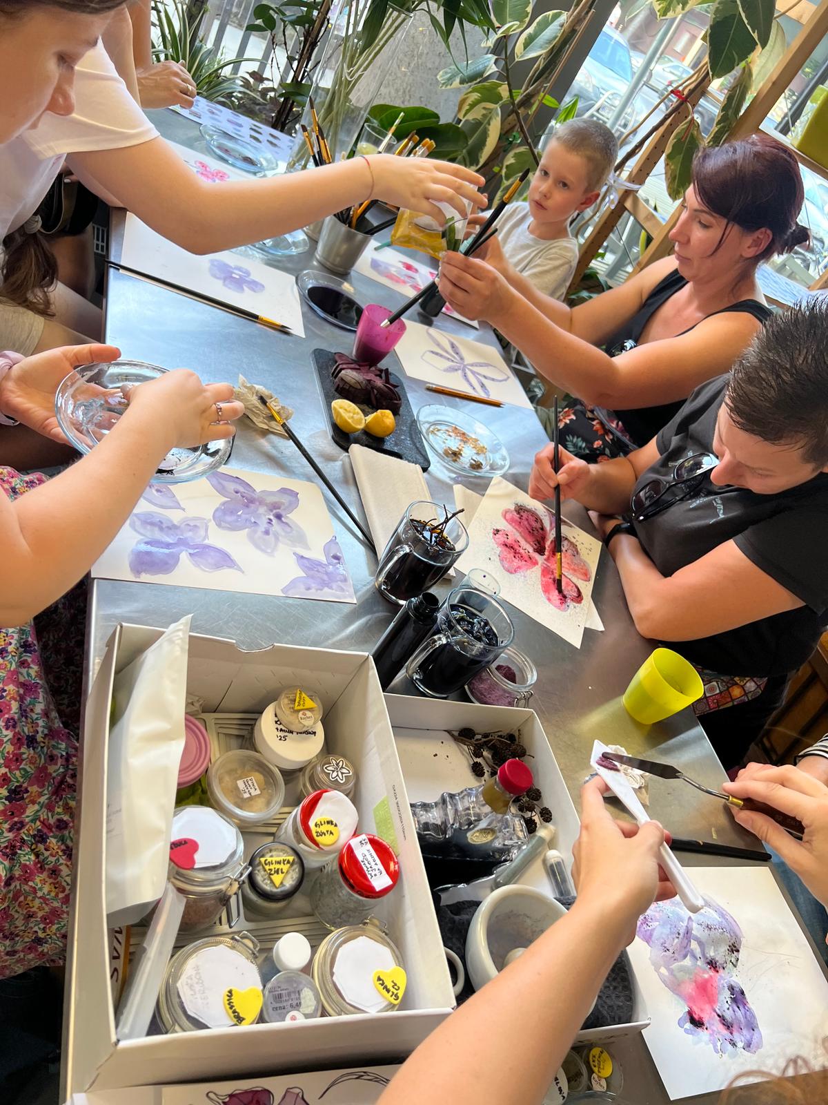 Grupa osób uczestniczy w warsztacie malarskim, tworząc kolorowe obrazy przy stole otoczonym roślinami.