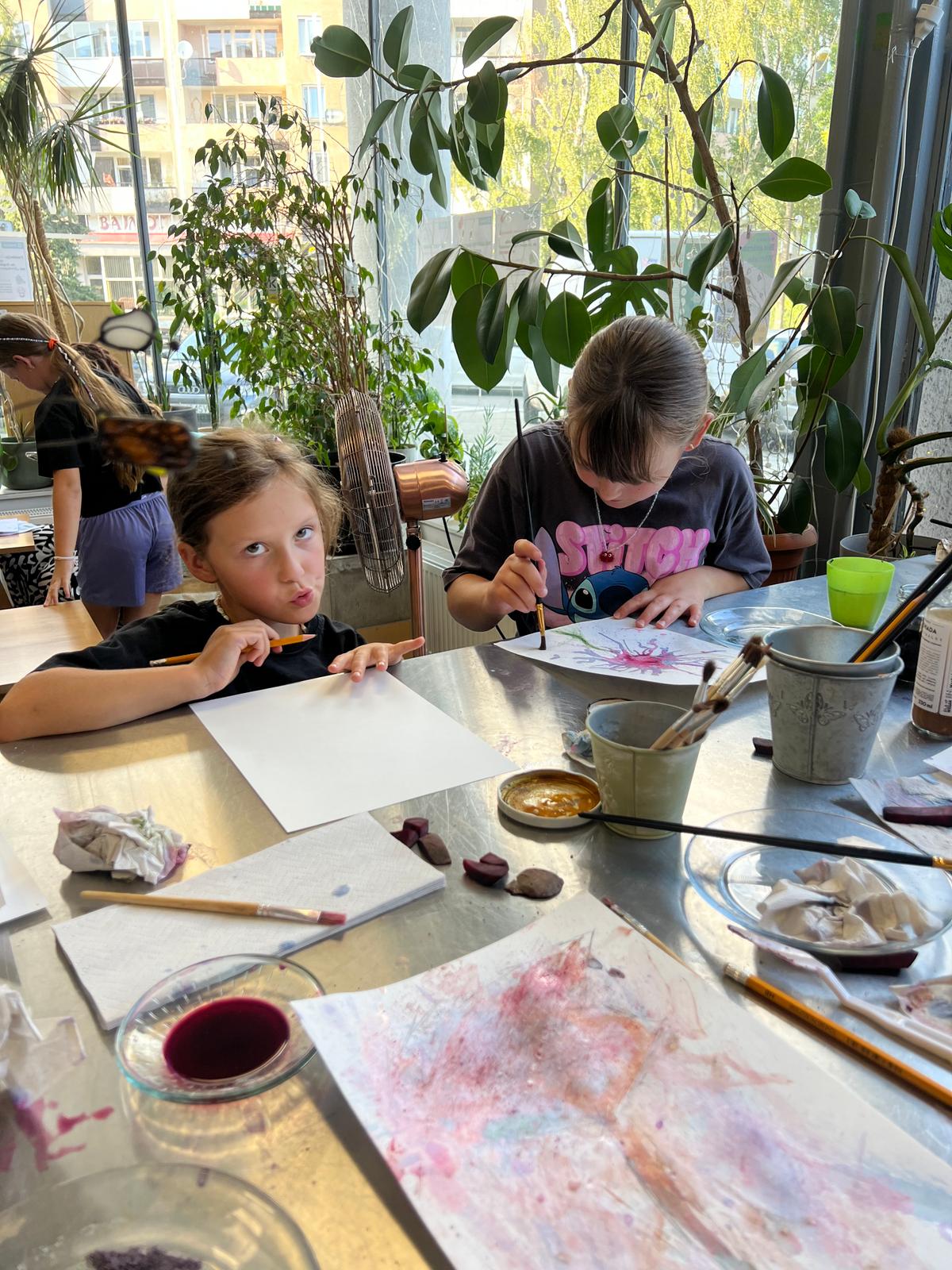 Dzieci malują akwarelą przy stole w jasnym pomieszczeniu pełnym zielonych roślin doniczkowych.
