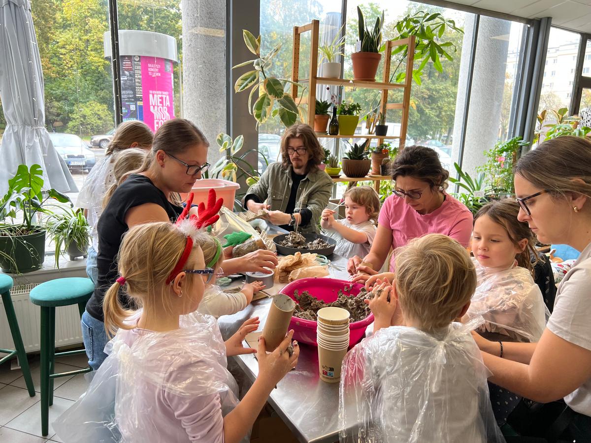 Grupa dzieci i dorosłych bierze udział w warsztacie sadzenia roślin w jasnym pomieszczeniu z oknami i roślinami doniczkowymi.