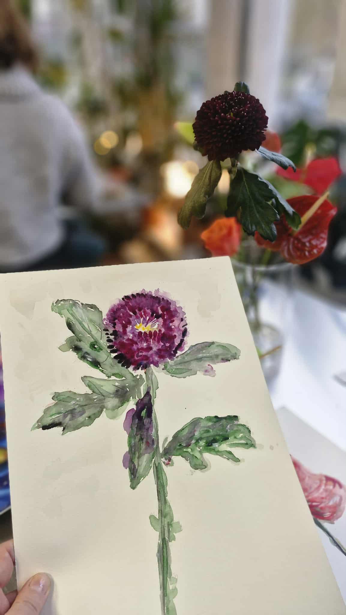 Akwarela ciemnoczerwonym kwiatem obok żywego kwiatu w wazonie na tle rozmytych ludzi i kwiatów.