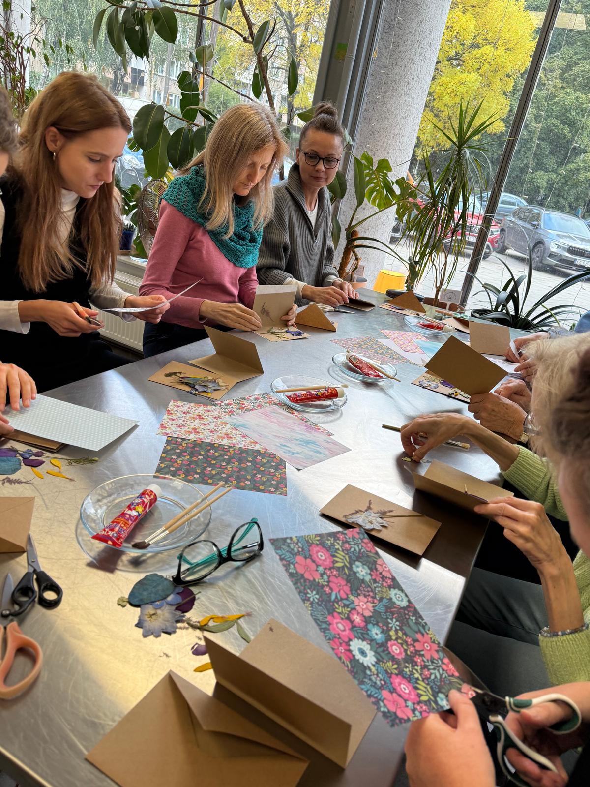 Grupa osób przy stole wykonuje pracę rękodzielniczą z papierem dekoracyjnym i pudełkami kartonowymi przy oknie.
