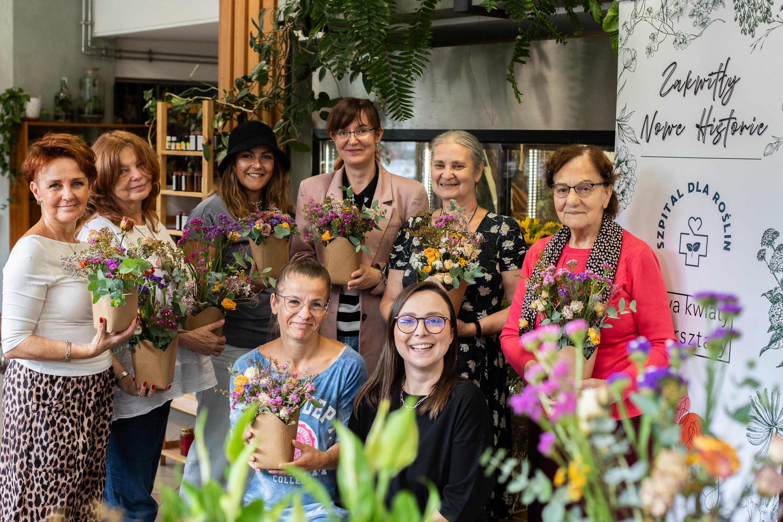 Grupa kobiet i dziewczyn trzymających kwiaty pozuje do zdjęcia w nowoczesnym wnętrzu pełnym roślin.