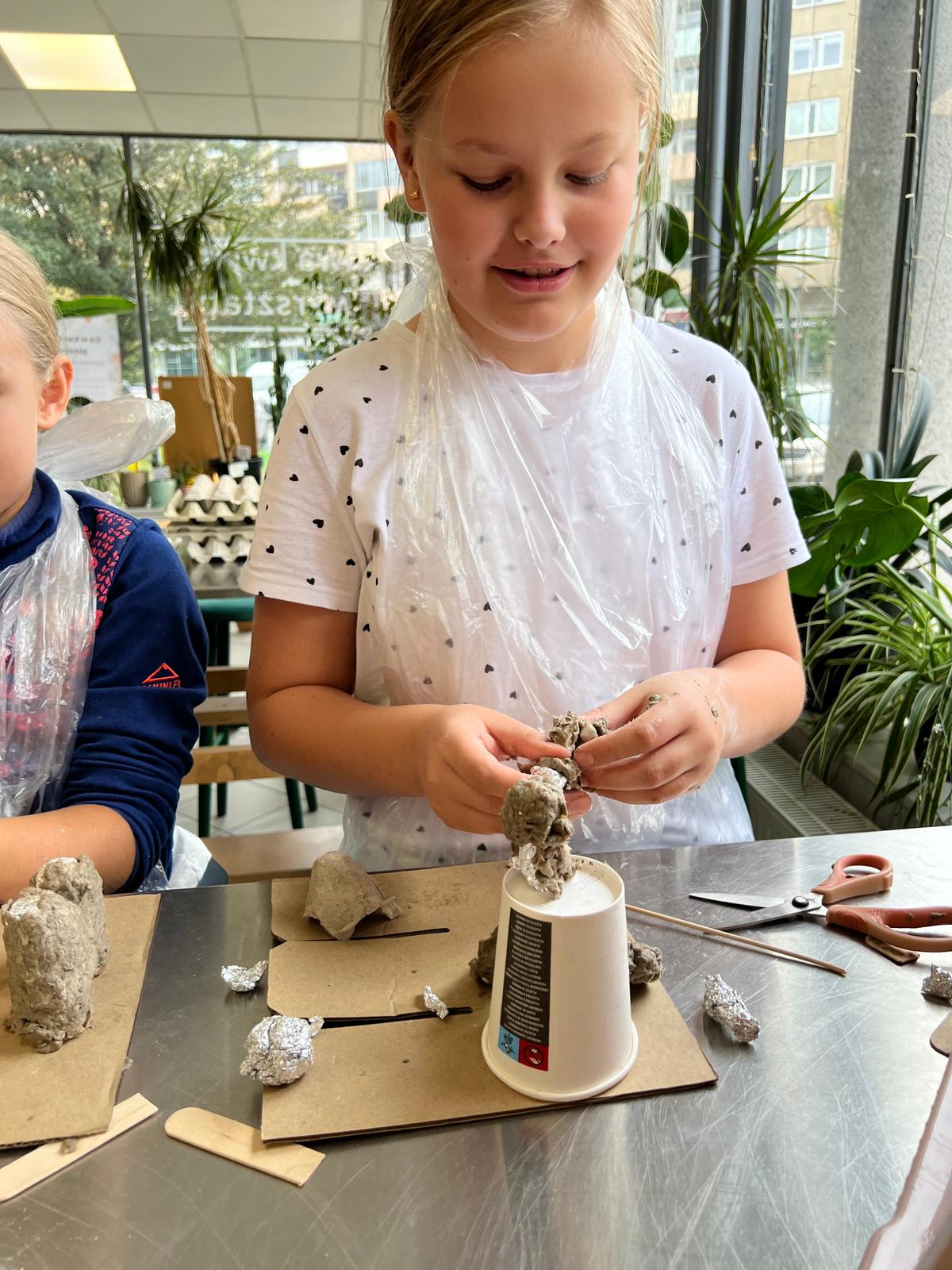 Dziewczynka w białej koszulce z serduszkami wykonuje eksperyment naukowy, mieszając materiały w pojemniku.