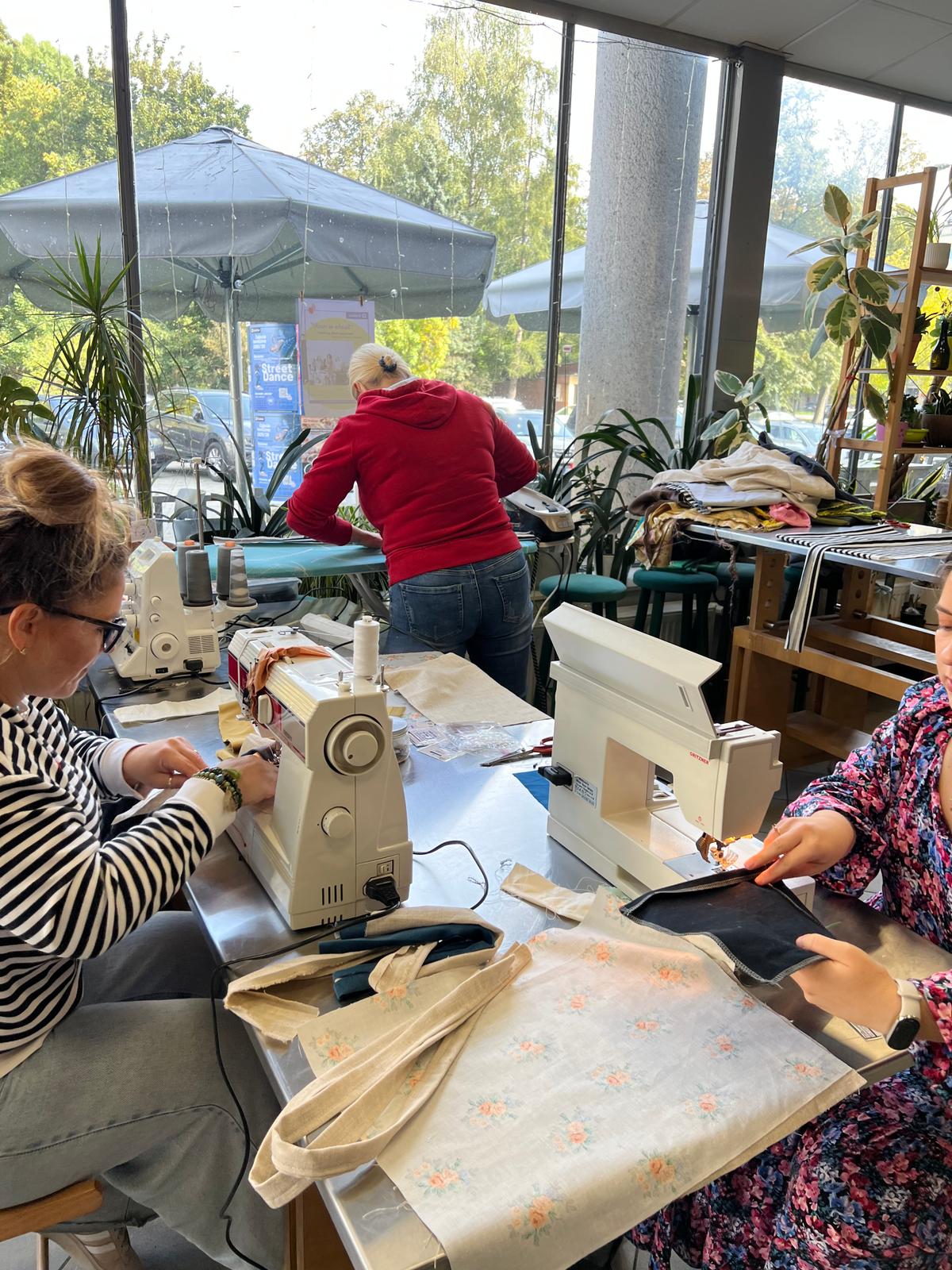 Trzy kobiety pracują przy maszynach do szycia w jasnym studio z dużymi oknami i roślinami doniczkowymi.