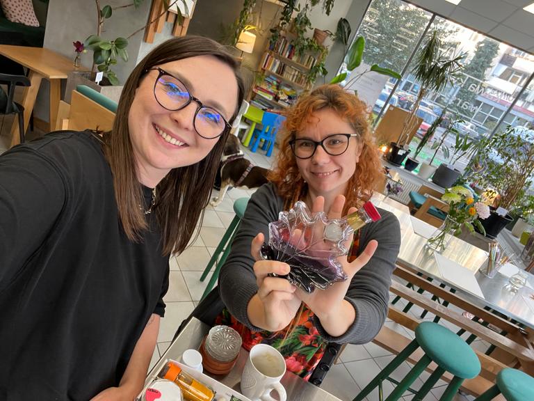 Dwie uśmiechnięte dziewczyny w kawiarni pokazują przezroczystą figurkę liścia, otoczone roślinami i książkami.
