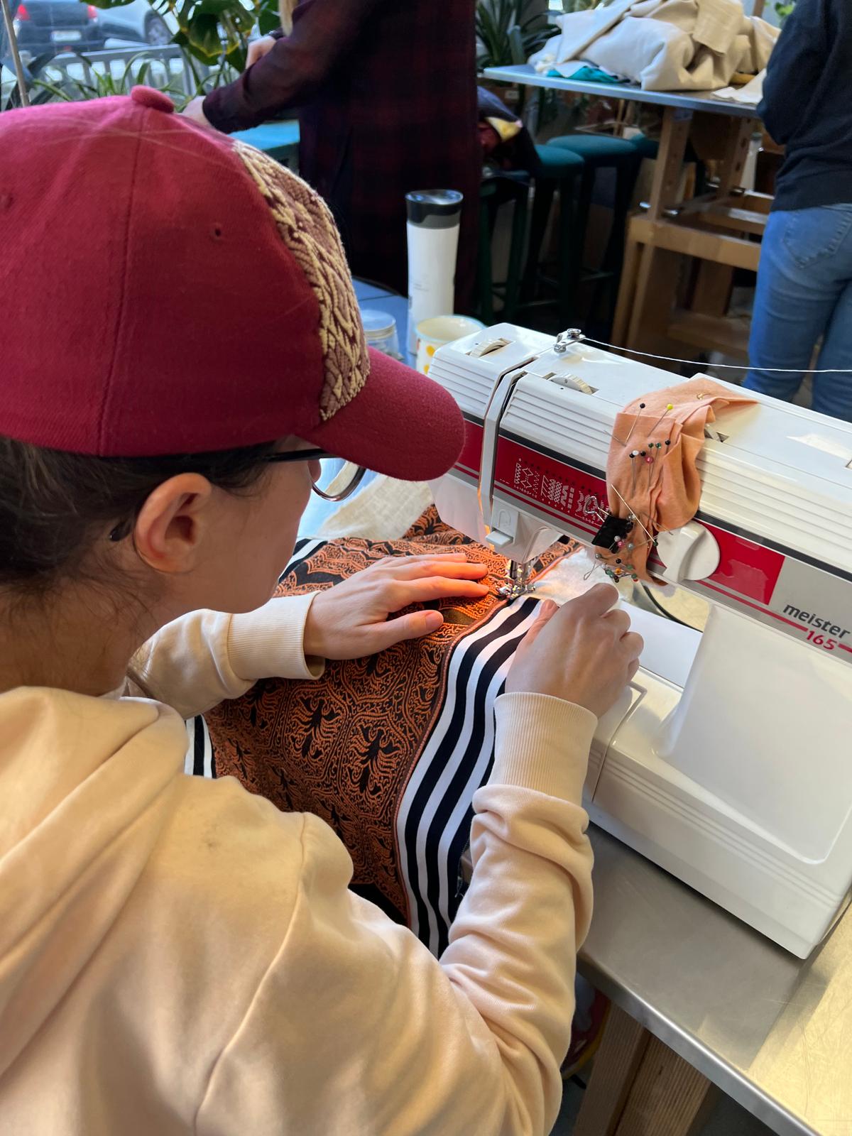 Dziewczynka w różowej czapce szyje na maszynie do szycia, pracując z brązową i czarno-białą tkaniną.