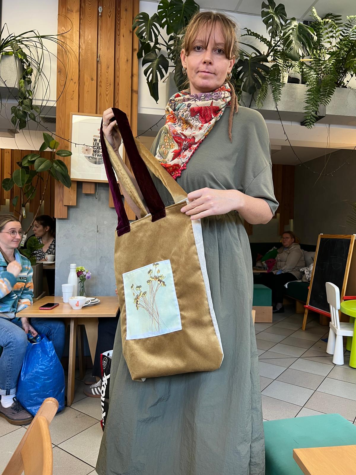 Kobieta prezentuje złotą torbę z botanicznym motywem na tle zielonych roślin w salonie.