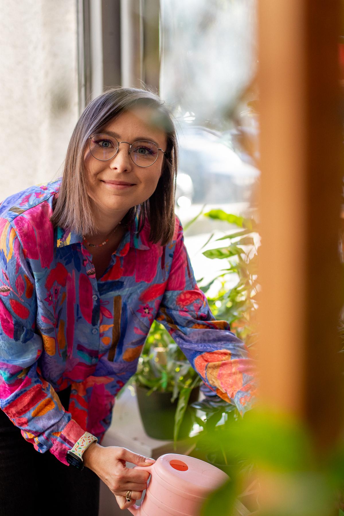 Kobieta w kolorowej koszuli i okularach opiekuje się roślinami na parapecie, trzymając różowy pojemnik.