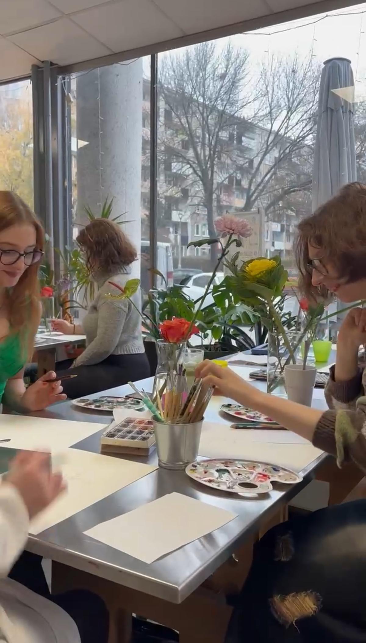 Grupa dzieci siedzi przy stole i maluje kwiaty, które stoją w wazonach przed dużymi oknami z widokiem na miasto.