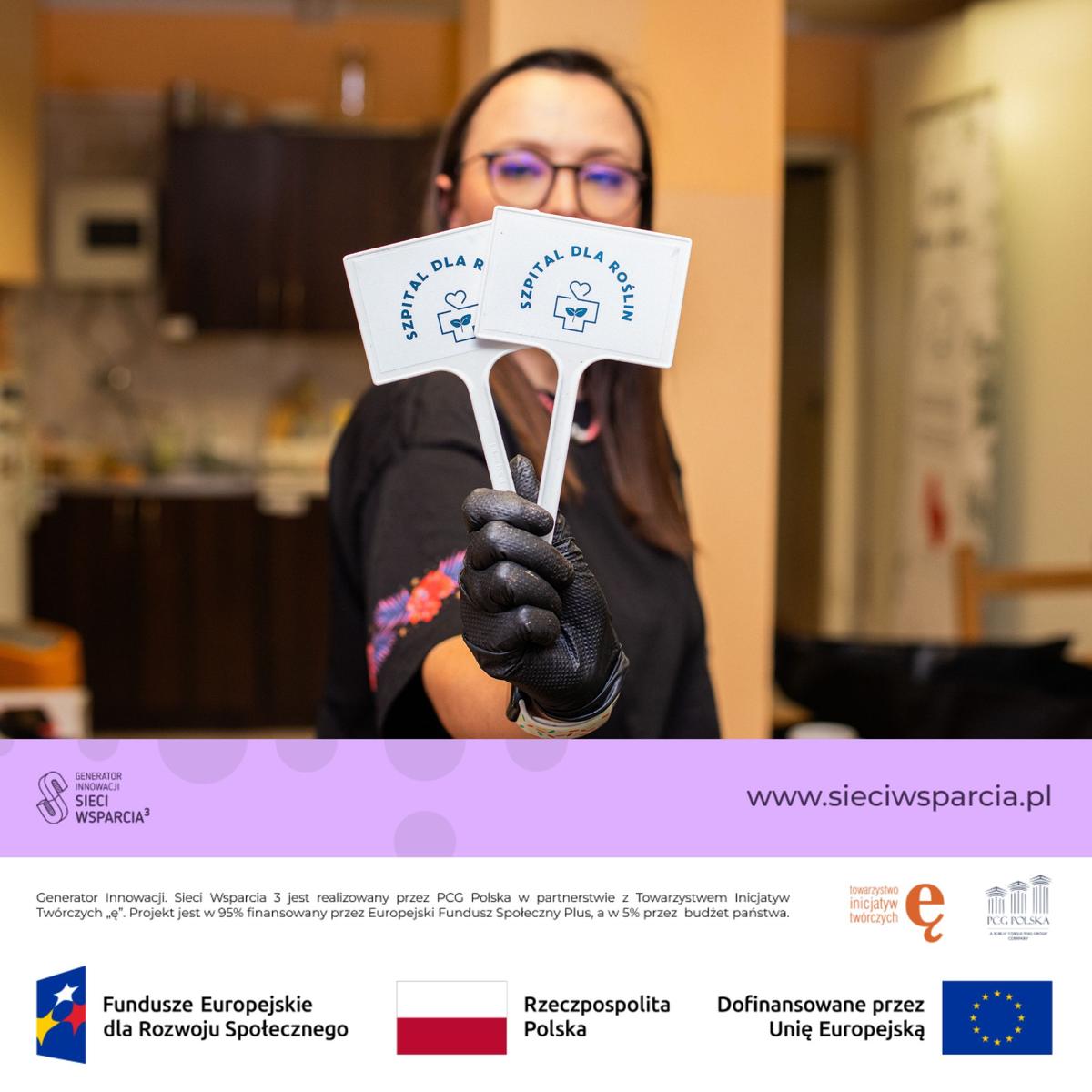 Osoba w czarnych rękawiczkach i okularach trzyma dwie białe kartki z napisem "Sprzedaj dla roślin" w kuchni.