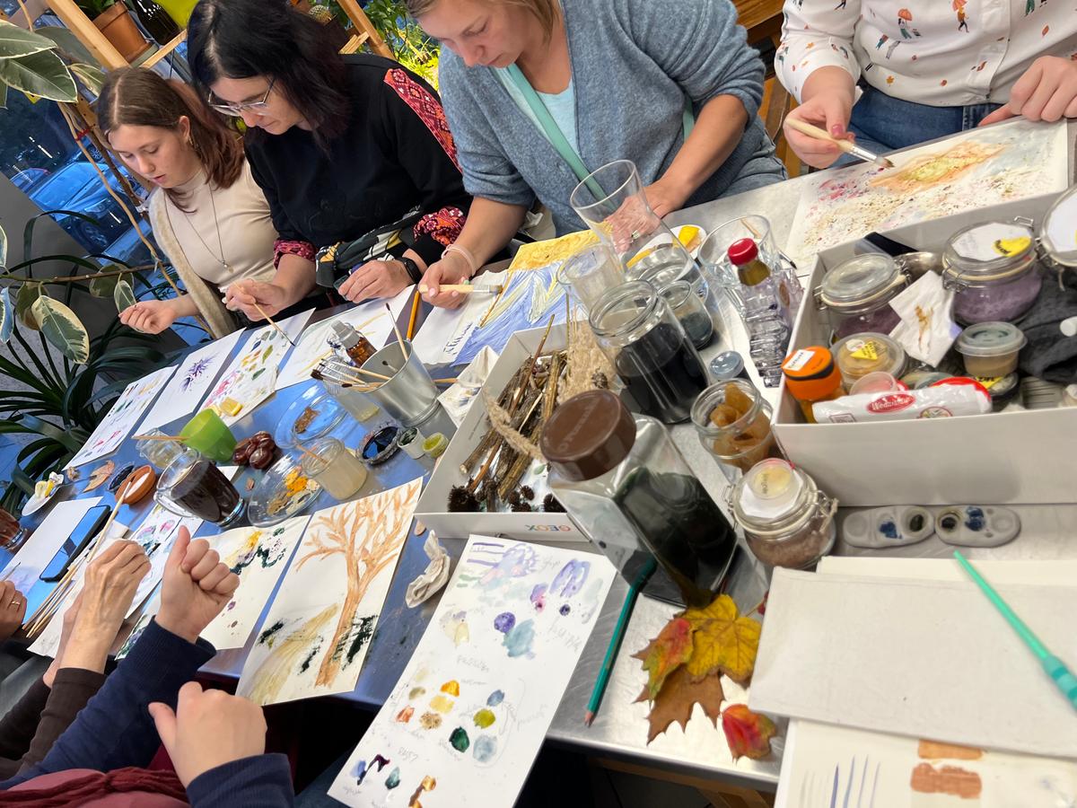 Grupa osób uczestniczy w warsztatach artystycznych, malując naturalne materiały oraz tworząc kolorowe prace przy stole.