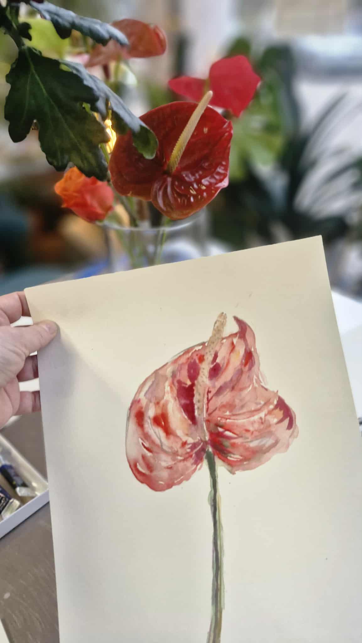 Osoba trzyma akwarelę przedstawiającą różowe kwiaty na tle rzeczywistych czerwonych kwiatów w wazonie.